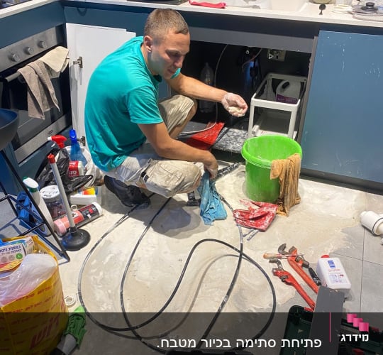 אינסטלטור עובד מתחת לכיור עם כלים ודלי ירוק
